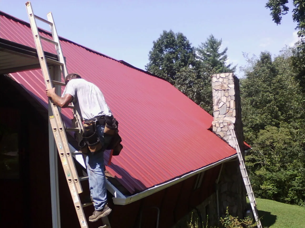 Roofer on a ladder beside a red metal roof and stone chimney — Soffit & Fascia Repair by Roofing Brothers in Wassaic